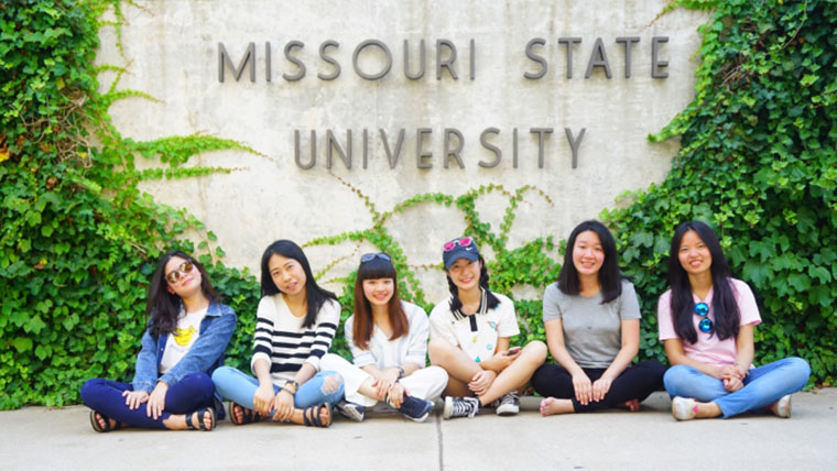 Group of students seating in front of a Missouri State University mural covered in ivy on a summer day.