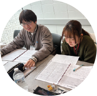 Two students sit at a table covered in notebooks and papers, focusing on their studies.