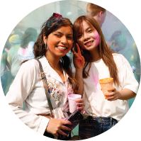 Two women covered in colorful Holi powder smiling and holding cups at an outdoor event.