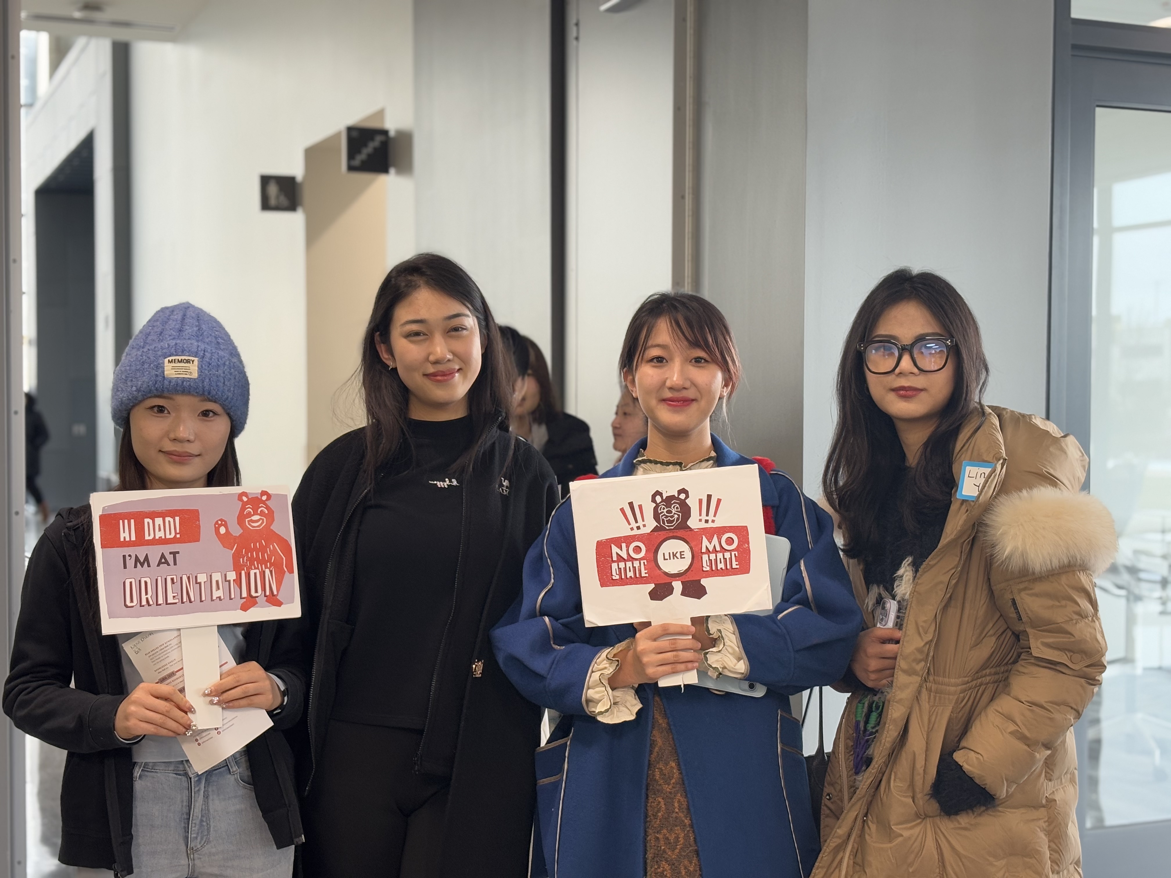 Four students standing indoors at an orientation event, holding signs and smiling at the camera.